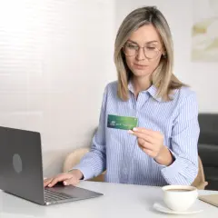 Kobieta z kartą płatniczą w ręku, gotowa do dokonania płatności online. Na biurku laptop, telefon i kawa. Reserved Paypo.
