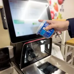 Ręka skanuje produkt przy kasie samoobsługowej. Ekran wyświetla informacje o zakupach i warunki płatności.