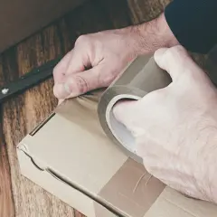 Ręce zaklejają taśmą kartonowe pudełko. Obok leżą nożyczki. Przygotowanie do wysyłki lub obi zwroty.
