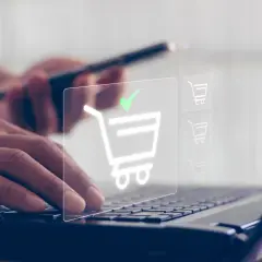 Dłonie wpisują dane na klawiaturze laptopa, a na ekranie pojawia się ikona koszyka z zielonym znacznikiem, symbolizująca udany zakup, jakby płatność przez paysafecard.