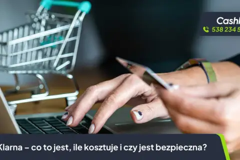 Dłoń trzyma kartę płatniczą nad klawiaturą laptopa, obok miniaturowy wózek sklepowy. Temat: karta Klarna.