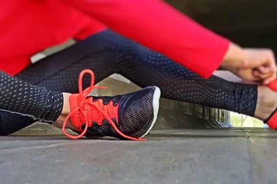 Osoba w czerwonej bluzie i czarnych legginsach z kropkami zawiązuje pomarańczowe sznurówki w butach sportowych. Idealne na martes sport zwroty.