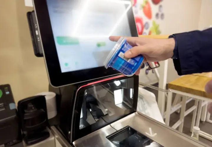 Ręka skanuje produkt przy kasie samoobsługowej. Ekran wyświetla informacje o zakupach i warunki płatności.