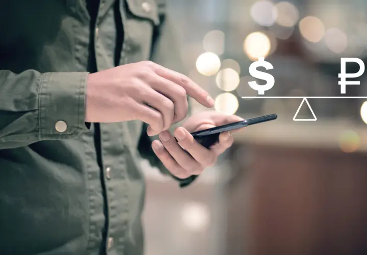 Dłoń trzyma telefon, na ekranie symbol dolara i rubla na wadze. Symbolizuje to paypal przewalutowanie.