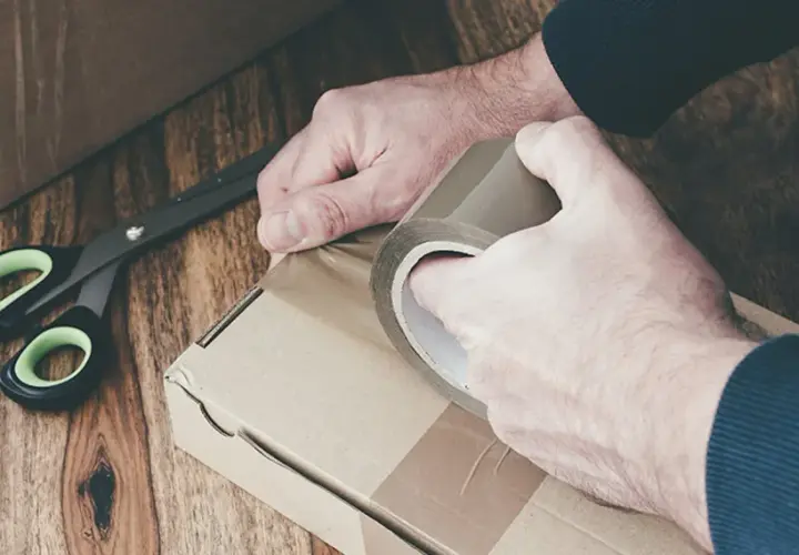 Ręce zaklejają taśmą kartonowe pudełko. Obok leżą nożyczki. Przygotowanie do wysyłki lub obi zwroty.
