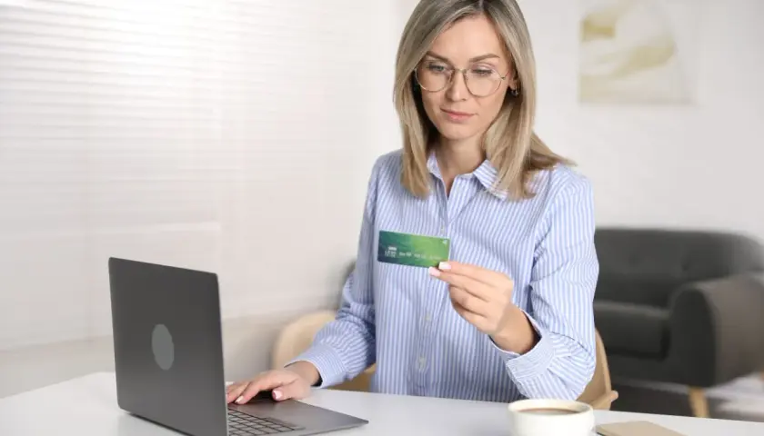Kobieta z kartą płatniczą w ręku, gotowa do dokonania płatności online. Na biurku laptop, telefon i kawa. Reserved Paypo.