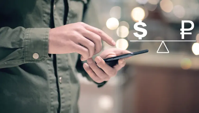 Dłoń trzyma telefon, na ekranie symbol dolara i rubla na wadze. Symbolizuje to paypal przewalutowanie.