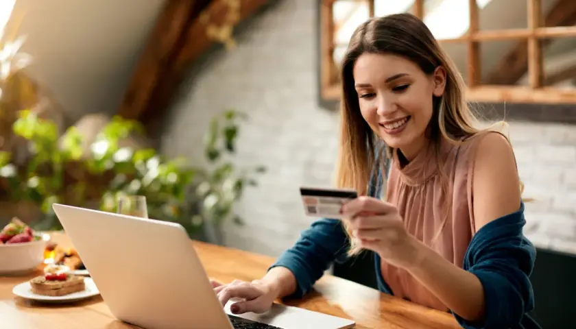 Uśmiechnięta kobieta z kartą płatniczą przy laptopie, planuje zakupy. Sprawdza cenę netto przed finalizacją transakcji.