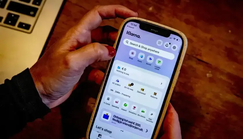 Dłoń trzyma smartfon z aplikacją Klarna. Czy Klarna jest bezpieczna? Aplikacja oferuje zakupy, płatności i zarządzanie zamówieniami.