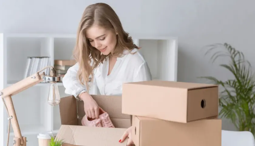 Kobieta rozpakowuje paczkę InPost, wyjmując z niej różowe buty. Obok stos kartonów.