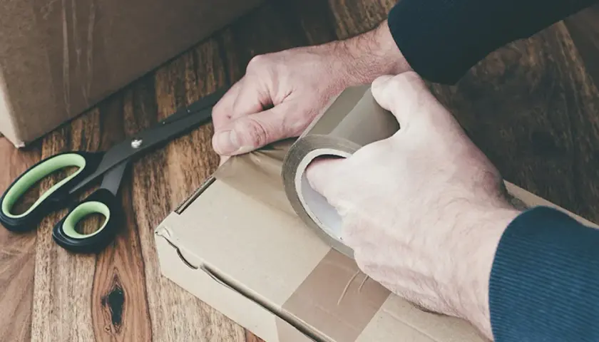 Ręce zaklejają taśmą kartonowe pudełko. Obok leżą nożyczki. Przygotowanie do wysyłki lub obi zwroty.