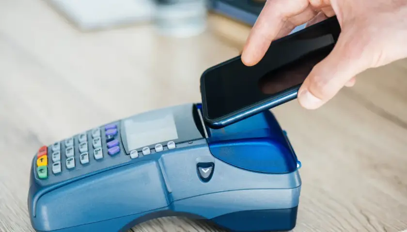 Dłoń zbliża smartfon do terminala płatniczego, realizując szybki booking paypo.