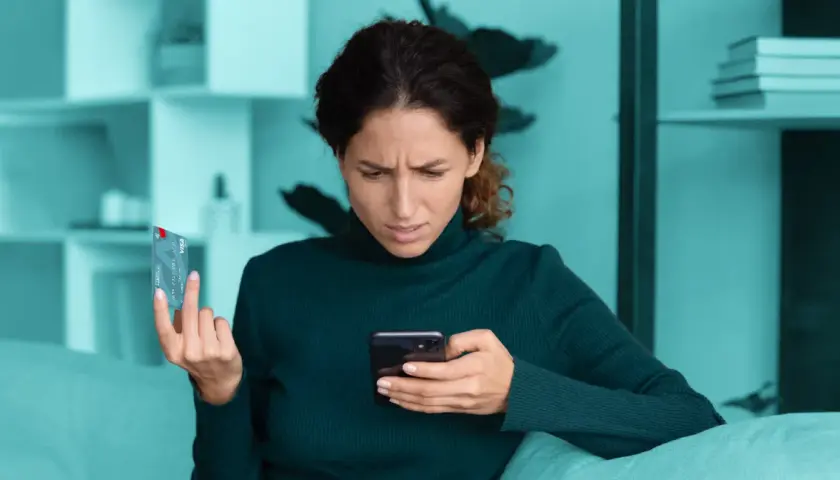 Zmarszczona kobieta trzyma kartę i telefon, zastanawiając się nad bezpieczne zakupy w internecie.