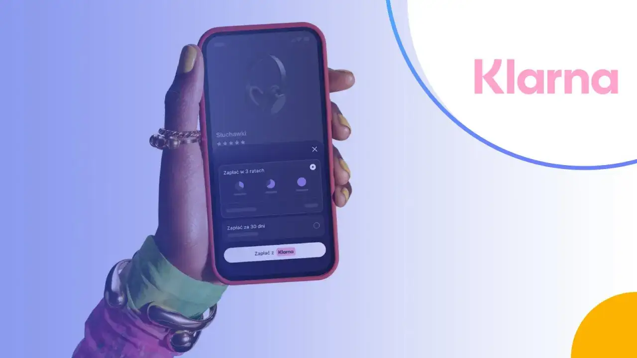 Dłoń trzyma telefon z aplikacją Klarna, oferującą płatność za słuchawki w ratach lub za 30 dni.