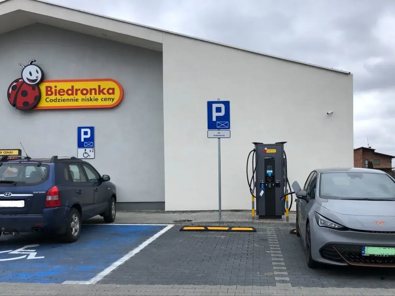 Dwa ekologiczne samochody ładują się przy Biedronce. Jeden to szary elektryk, drugi to niebieski SUV.